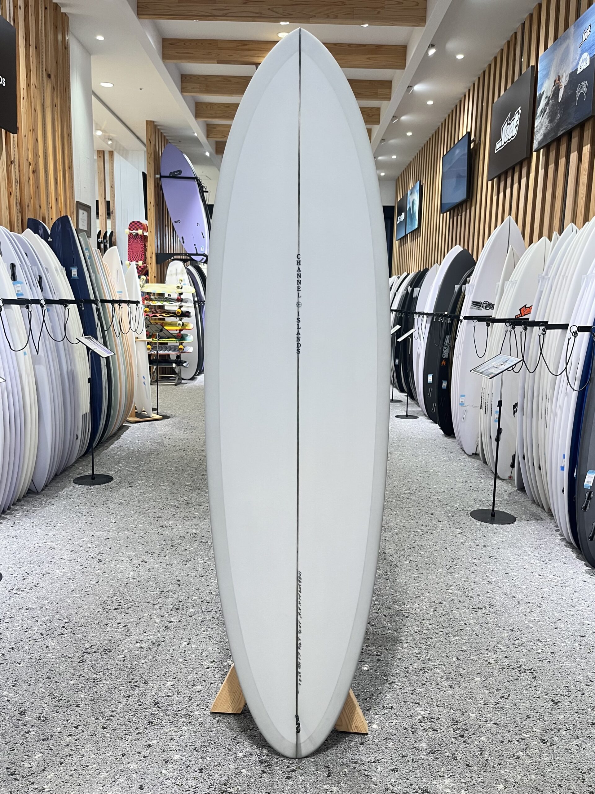 IMG_2673 - SHOP NEWS | ムラサキスポーツオンラインストア 通販 Tall white surfboard on display in a showroom, surrounded by racks of assorted surfboards along wooden-paneled walls.