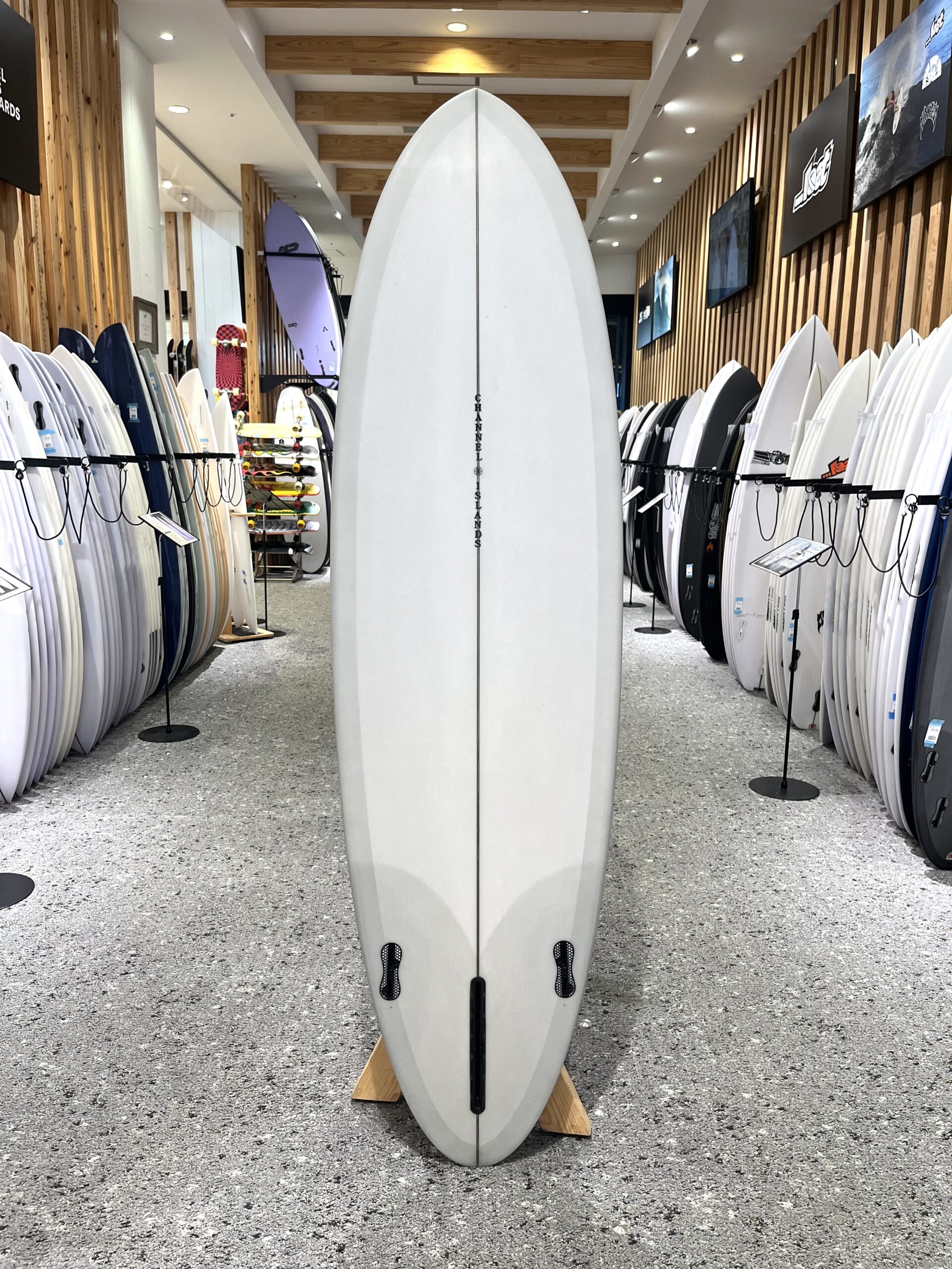 IMG_2678 - SHOP NEWS | ムラサキスポーツオンラインストア 通販 Tall white surfboard standing upright on a wooden stand in a surf shop, flanked by racks of boards on both sides and wood-paneled walls.