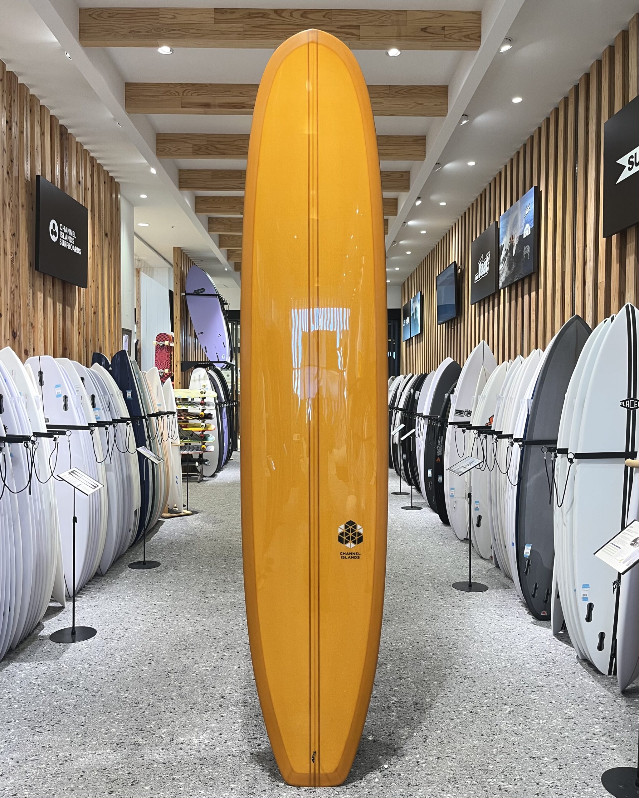 IMG_2681 - SHOP NEWS | ムラサキスポーツオンラインストア 通販 Tall orange surfboard stands upright in a showroom aisle, with rows of white surfboards on both sides and wooden slat walls behind.