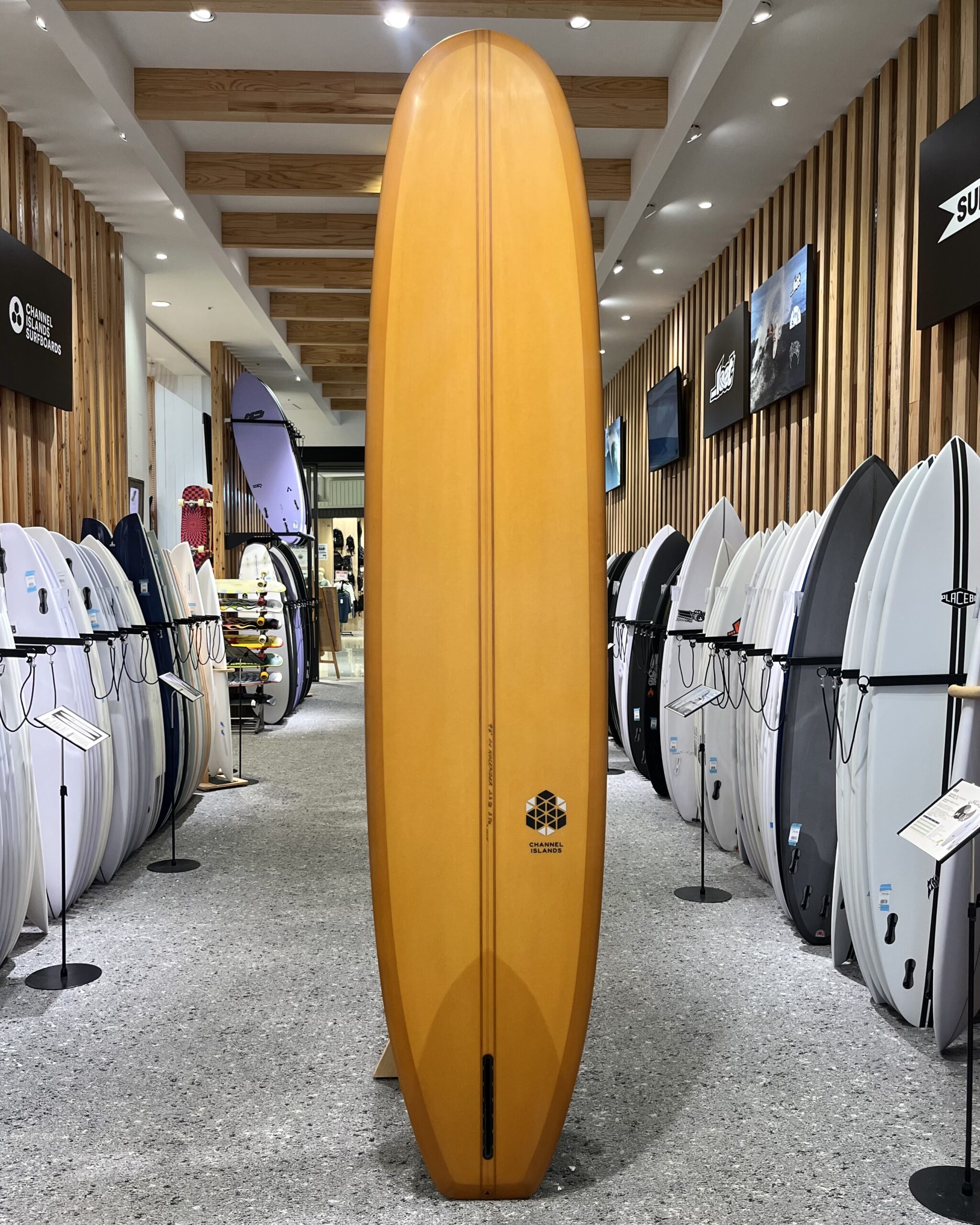 IMG_2693 - SHOP NEWS | ムラサキスポーツオンラインストア 通販 Tall orange surfboard standing upright in a surf shop between rows of white and gray boards on racks.