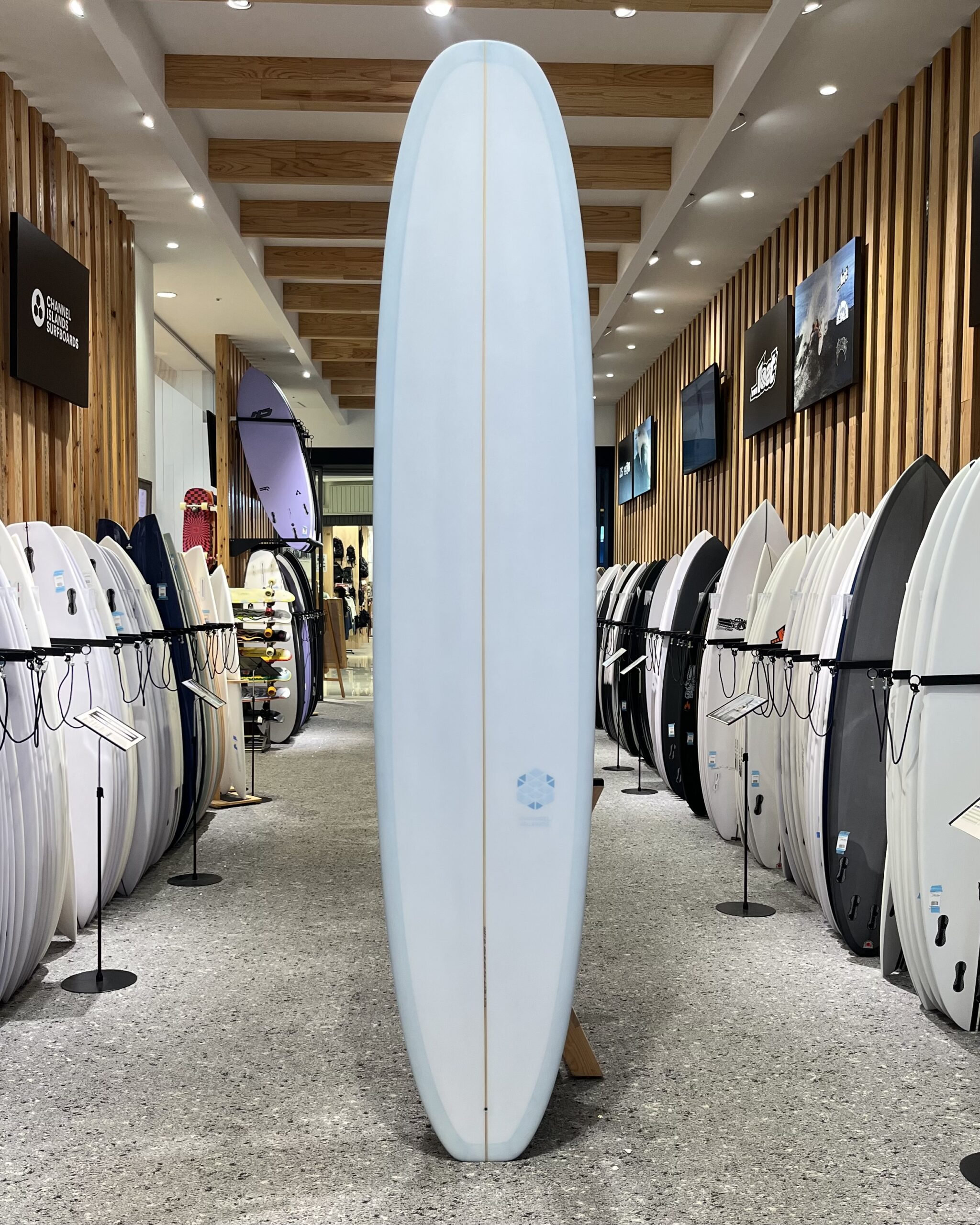 IMG_2694 - SHOP NEWS | ムラサキスポーツオンラインストア 通販 Tall light-blue surfboard displayed upright in a store aisle with rows of boards on both sides.