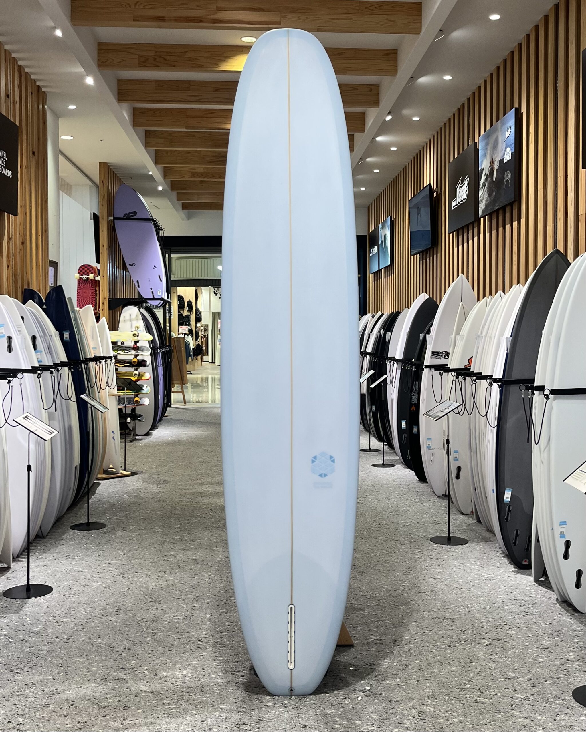 IMG_2704 - SHOP NEWS | ムラサキスポーツオンラインストア 通販 Tall light-blue surfboard stands upright in a shop aisle, flanked by rows of smaller boards along wooden slat walls.