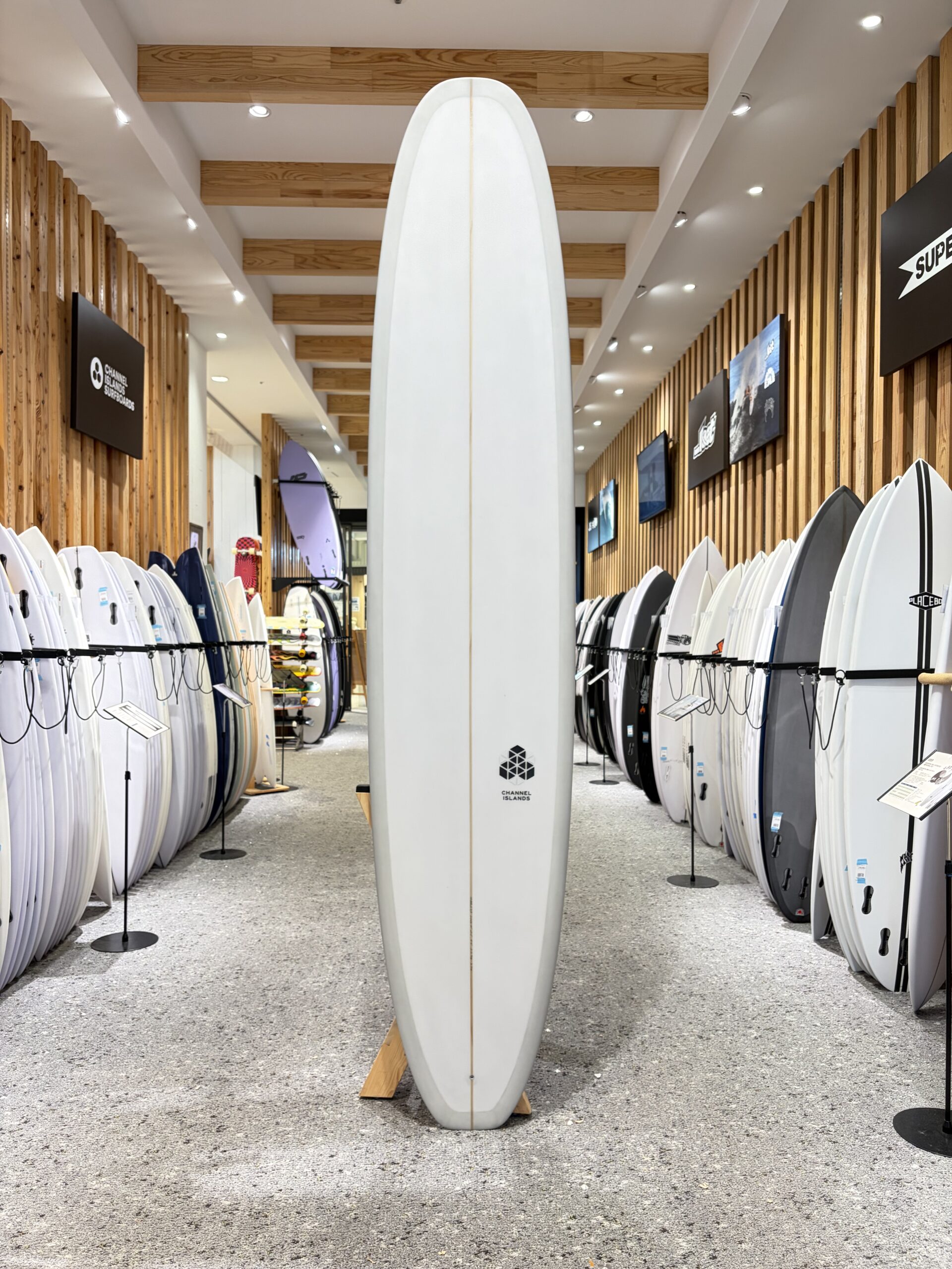 IMG_4531 - SHOP NEWS | ムラサキスポーツオンラインストア 通販 Tall white surfboard standing upright in a showroom with rows of boards along wooden walls and bright ceiling lights.
