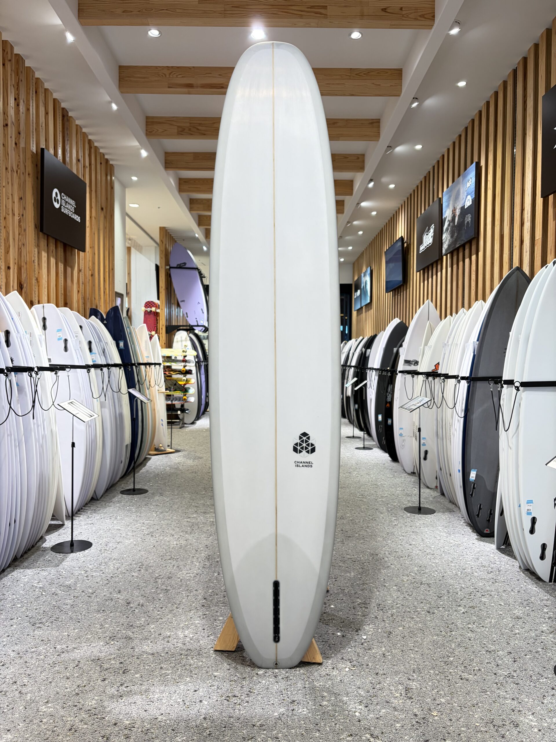 IMG_4532 - SHOP NEWS | ムラサキスポーツオンラインストア 通販 Tall white surfboard standing upright in the center of a showroom with numerous boards lined along walls on both sides, wooden beams above.
