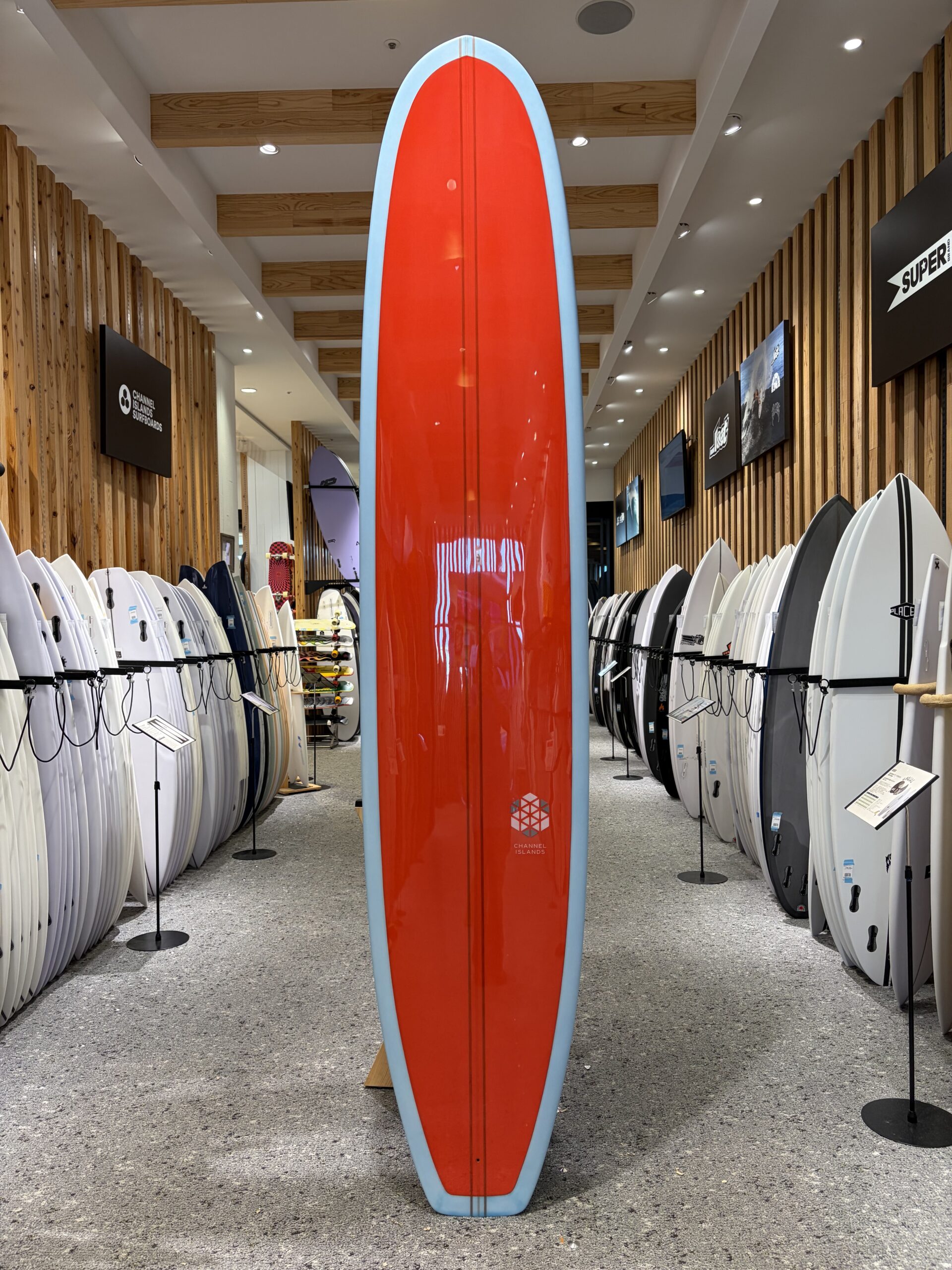 IMG_4533 - SHOP NEWS | ムラサキスポーツオンラインストア 通販 Tall red surfboard with light blue edges stands upright in a surf shop display, flanked by white boards on both sides.