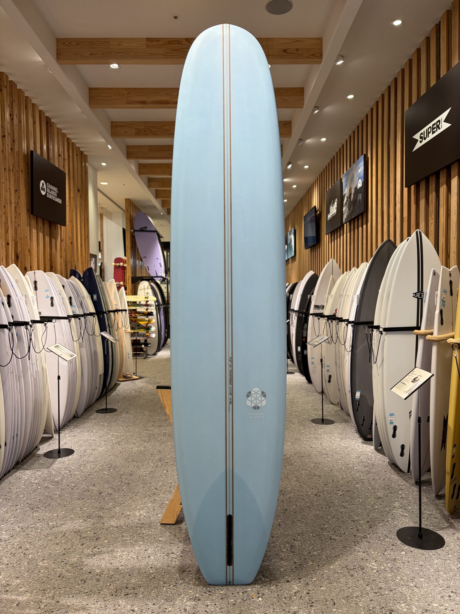 IMG_4535 - SHOP NEWS | ムラサキスポーツオンラインストア 通販 Tall light-blue surfboard stands upright as a centerpiece between rows of white surfboards in a showroom.