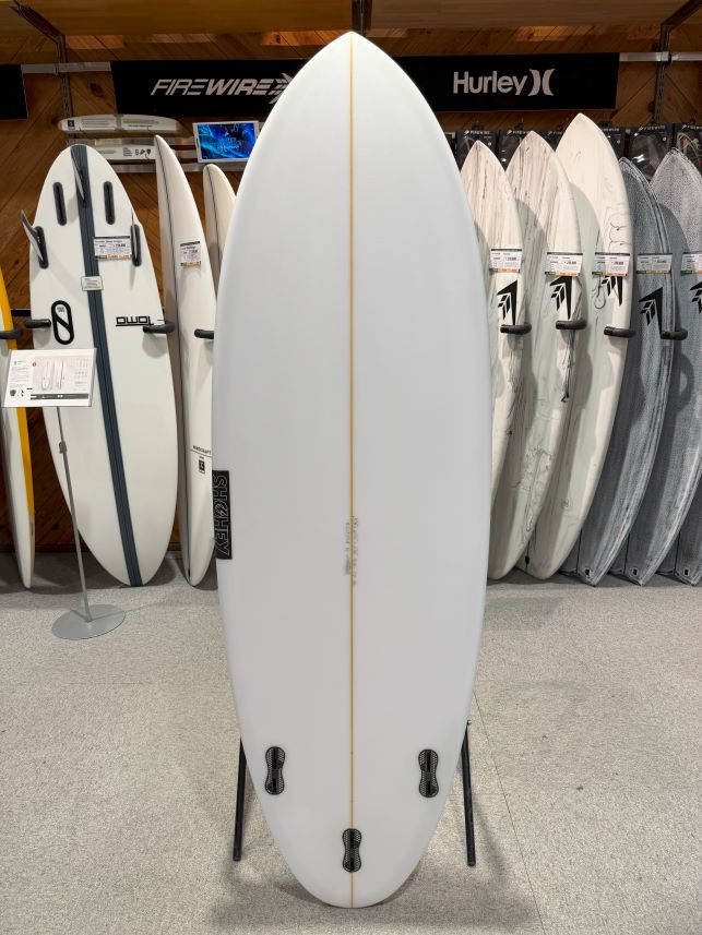White pointed surfboard on a stand in a showroom, with other boards lined up behind it and wooden walls in the background.