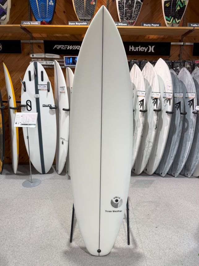 IMG_8788 - SHOP NEWS | ムラサキスポーツオンラインストア 通販 Tall white surfboard on a stand in a shop, with multiple boards on racks behind it and brand signs above (Firewire, Hurley).