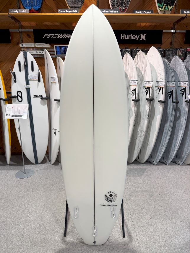 IMG_8789 - SHOP NEWS | ムラサキスポーツオンラインストア 通販 Light gray surfboard standing upright on a floor rack in a shop, with other boards lined up behind it; features a central seam and the VELL Three Weather logo near the tail.