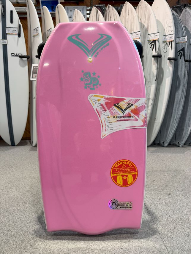 Pink bodyboard standing upright on a showroom floor, with teal graphics at the top and a warning sticker on the lower half, surrounded by other boards in the store.