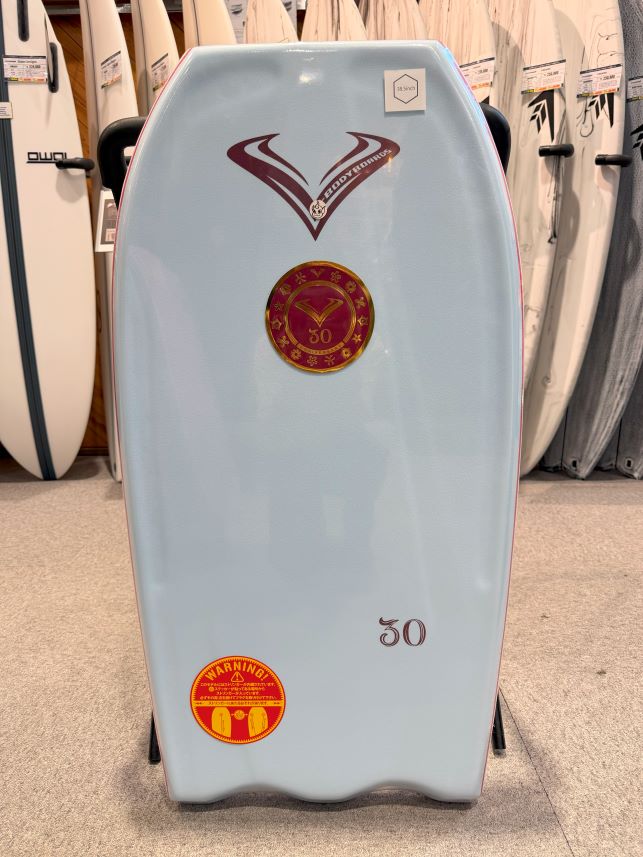 Light blue surfboard standing upright in a shop, featuring red and gold logos and the number 30, among other boards in the background.