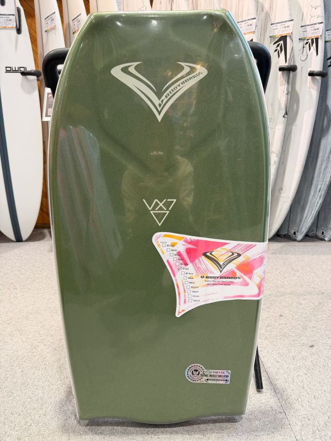 Green surfboard on display with V-shaped logo near top and a colorful sheet attached to lower right side in a shop window.