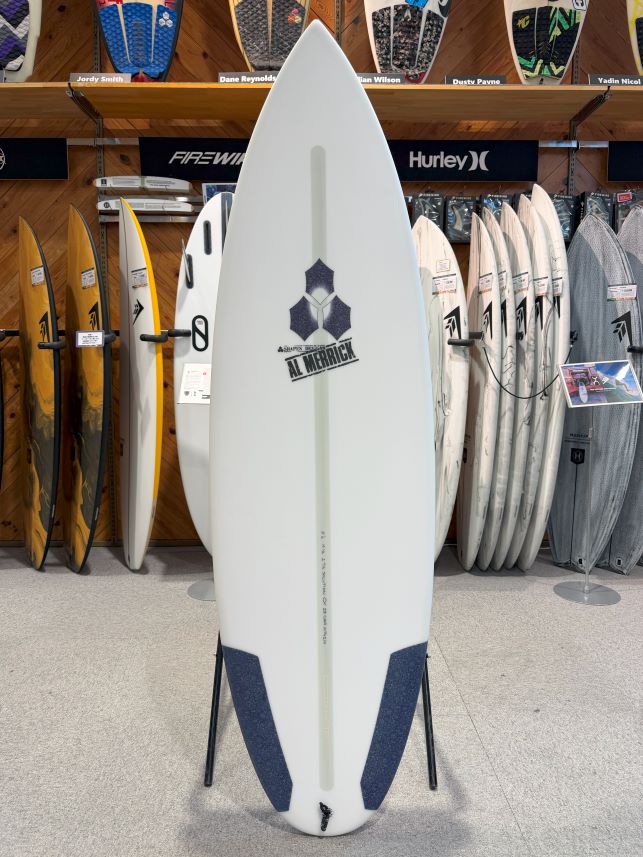 White surfboard on a stand in a surf shop, logo visible along the center, with other boards on racks in the background.
