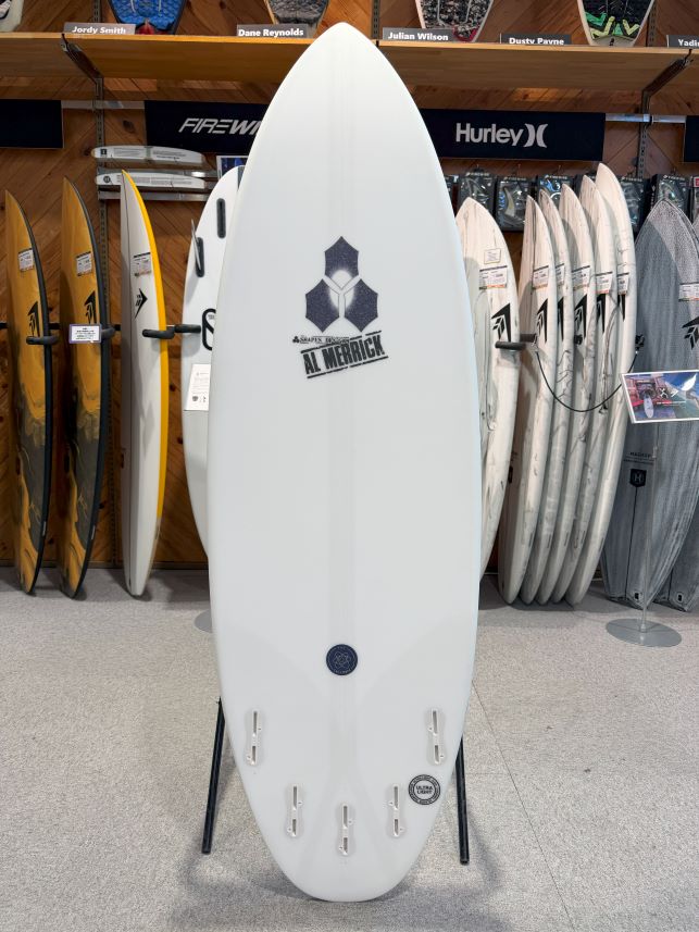 White surfboard on a stand in a surf shop, prominently displaying the Al Merrick logo with other boards on racks in the background.