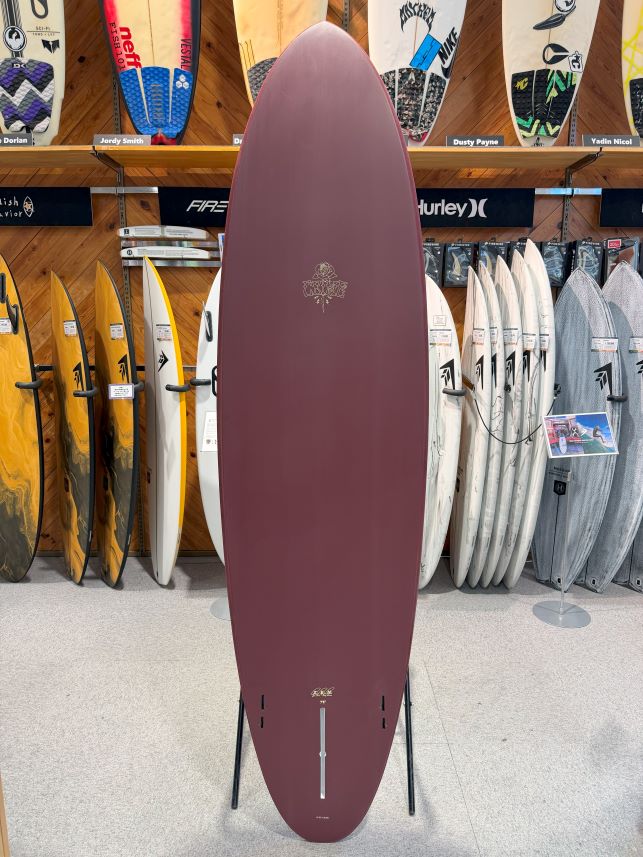 Maroon surfboard standing upright on a stand in a store, surrounded by other boards on racks behind it.