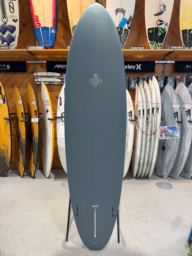 Gray surfboard on a stand in a surf shop, with colorful boards on racks in the background.