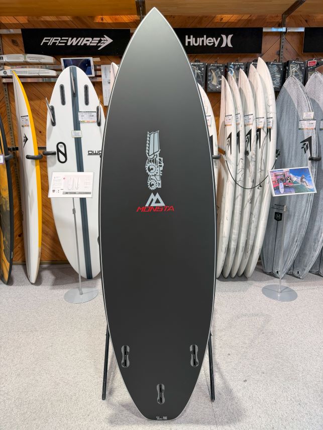 IMG_9221 - SHOP NEWS | ムラサキスポーツオンラインストア 通販 Black Monsta surfboard on a display stand in a shop, featuring a rocket/gear graphic; rows of white and gray boards behind it.
