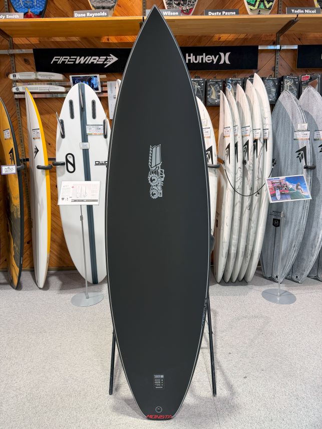 IMG_9222 - SHOP NEWS | ムラサキスポーツオンラインストア 通販 Dark gray surfboard standing upright on a stand in a surf shop, with other boards on racks behind it.