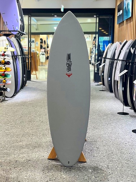 Gray surfboard on a wooden stand in a surf shop, surrounded by other boards on racks inside a showroom.
