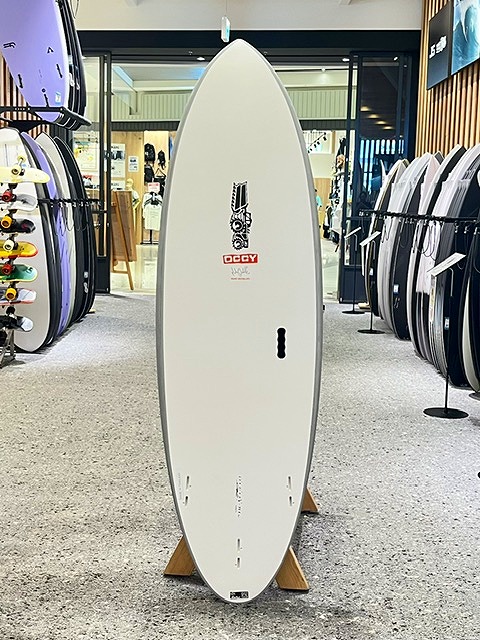 White surfboard standing upright on a wooden rack inside a surf shop, surrounded by other boards on racks.