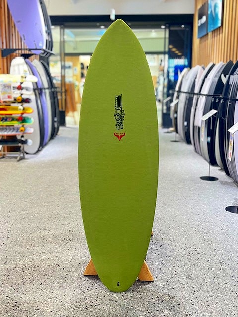Green longboard surfboard standing upright on a wooden rack inside a surf shop, with other boards visible on shelves around it.
