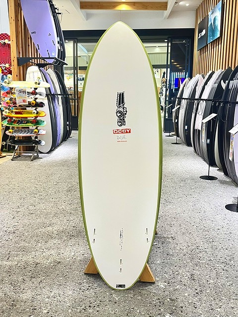 White surfboard with a lime-green edge on a wooden display stand in a surf shop, surrounded by other boards on racks.