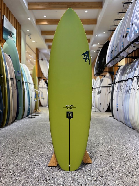 a-image0 - SHOP NEWS | ムラサキスポーツオンラインストア 通販 Lime-green surfboard standing upright on a display stand in a surf shop, with other boards lining the walls.