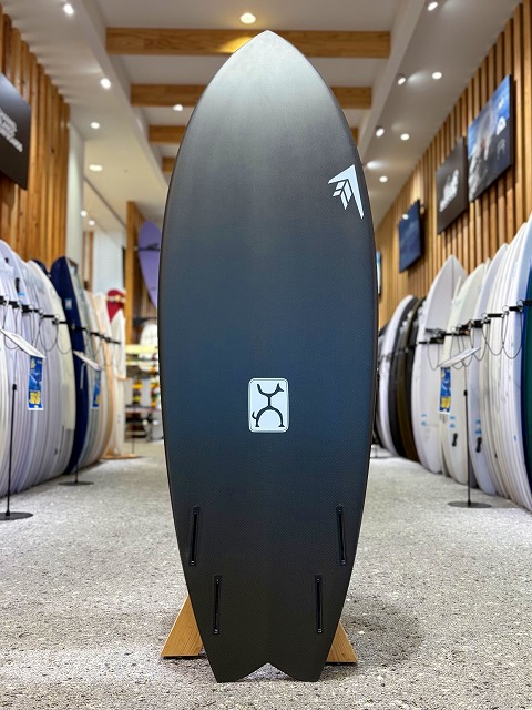 Dark gray surfboard on a wooden stand in a surf shop, with two fin boxes at the tail and white logos near the top.
