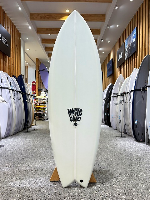 White surfboard standing upright on a wooden rack in a surf shop, with wood-paneled walls and many other boards on either side, featuring a black 'Magic' logo.