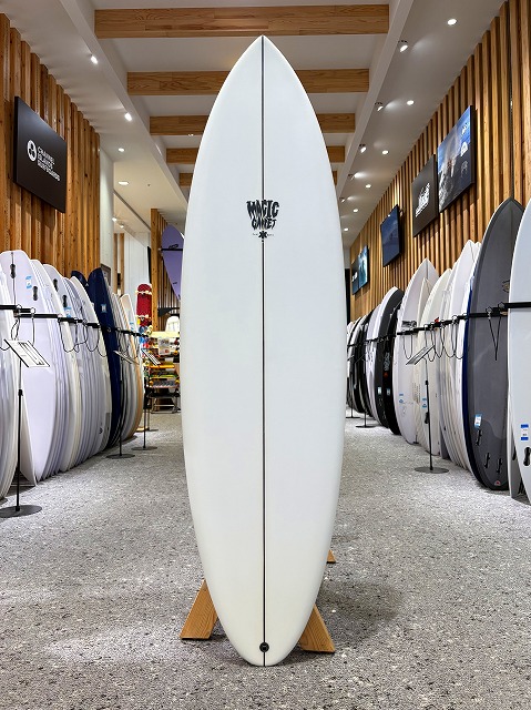 White surfboard standing upright on a wooden rack in a showroom, surrounded by other boards and wall-mounted displays in a surfing shop.