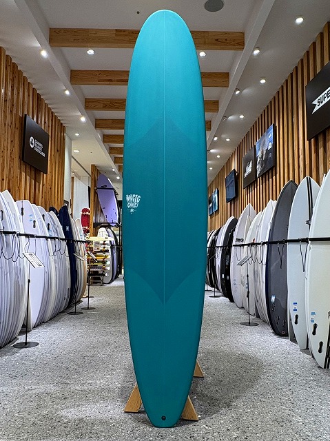 Tall turquoise surfboard standing upright on a wooden display stand inside a surf shop, surrounded by racks of other boards along the walls.