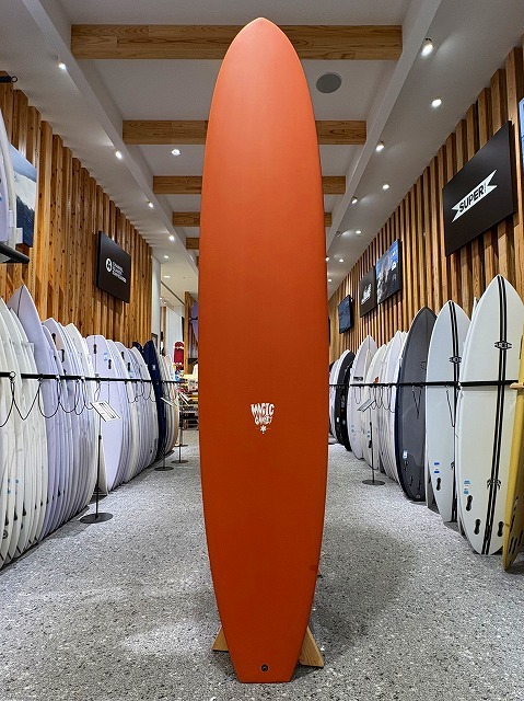 Orange surfboard standing upright on a showroom floor between rows of white and light-colored surfboards, with wooden wall panels and overhead lights in a surf shop.
