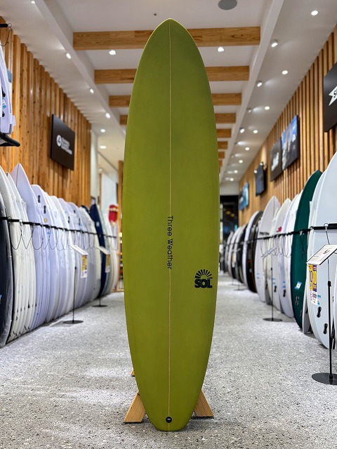 Tall green longboard surfboard on a wooden stand in a surf shop aisle, with other boards lined up on both sides and 'three watermen' text plus an SOL logo on the board.