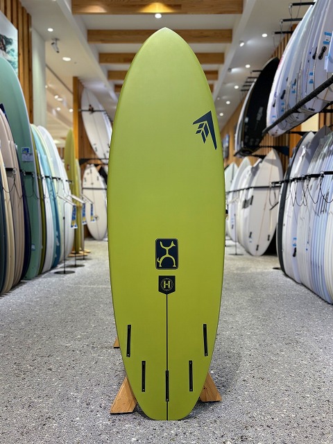 a-image1 - SHOP NEWS | ムラサキスポーツオンラインストア 通販 Yellow-green surfboard standing upright in a surf shop, with other boards on racks along the aisle behind it.