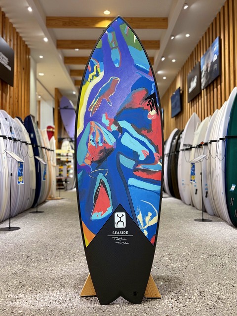 Colorful abstract-art surfboard on a stand in a surf shop, with rows of boards along the aisle.
