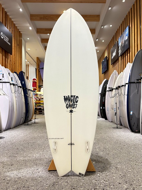 White surfboard with a 'Magic' logo standing upright on a wooden stand in a surf shop showroom. Surrounding racks hold numerous boards on both sides.