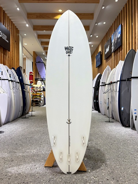 White surfboard standing upright on a display rack in a surf shop, with other boards lined up along the walls and wood-paneled ceiling in the background.