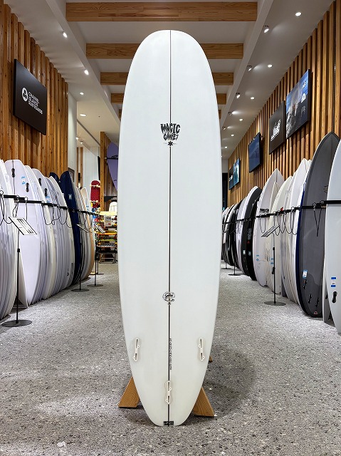 White surfboard upright on a wooden stand in a surf shop, with rows of other boards on racks along wood-paneled walls.