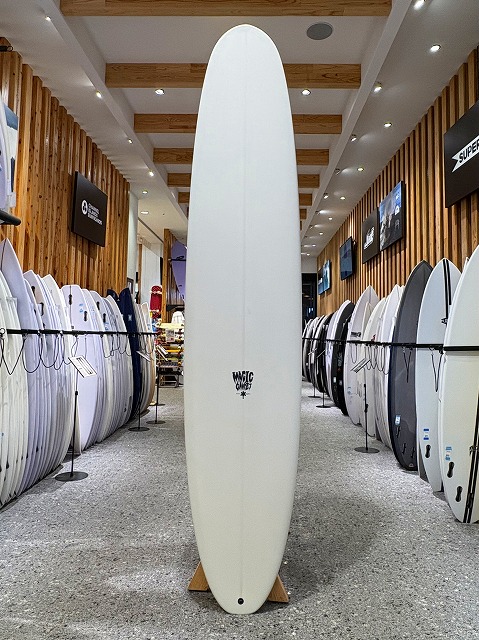 White longboard surfboard standing upright on a wooden display stand in a surf shop, flanked by many other surfboards along racks.