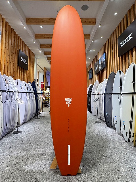 Tall orange surfboard displayed upright on a stand in a surf shop, flanked by white and colored boards along wooden walls.
