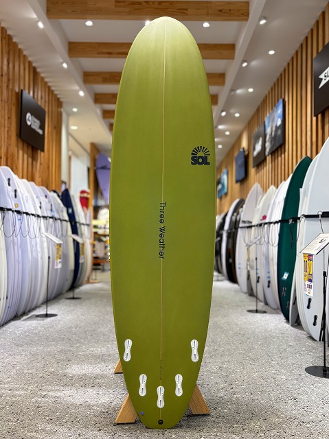 Lime green longboard surfboard on display in a surf shop, standing upright on a stand with racks of boards on both sides.