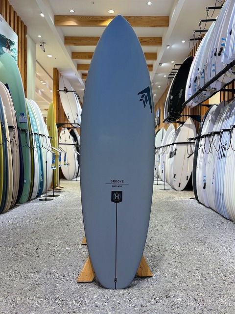 a-image2 - SHOP NEWS | ムラサキスポーツオンラインストア 通販 Blue surfboard standing upright on a wooden rack in a surf shop, with rows of colorful boards lining both sides of the aisle.