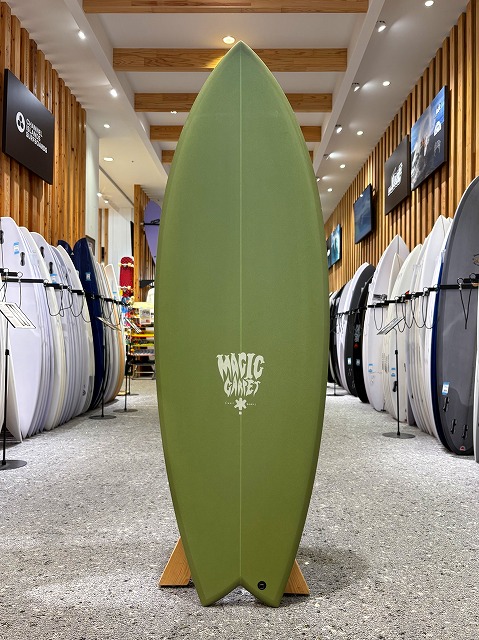 Green surfboard on a wooden stand in a surf shop showroom, centered with the 'Magic' logo, surrounded by rows of other boards on racks.