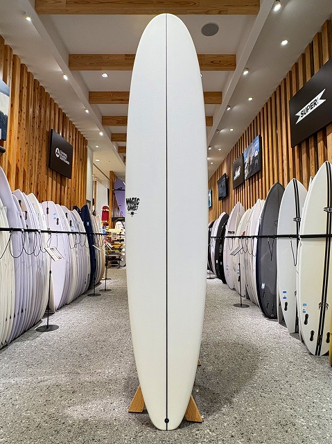 Tall white surfboard standing upright on a wooden stand in a surf shop showroom amid rows of boards on both sides.