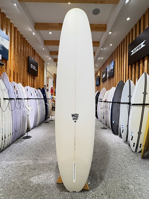 Tall white surfboard displayed upright in a surf shop, flanked by racks of boards along wooden walls and bright overhead lighting.
