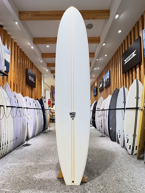 White surfboard standing upright on a wooden rack in a surf shop aisle, with numerous boards lined along both walls.