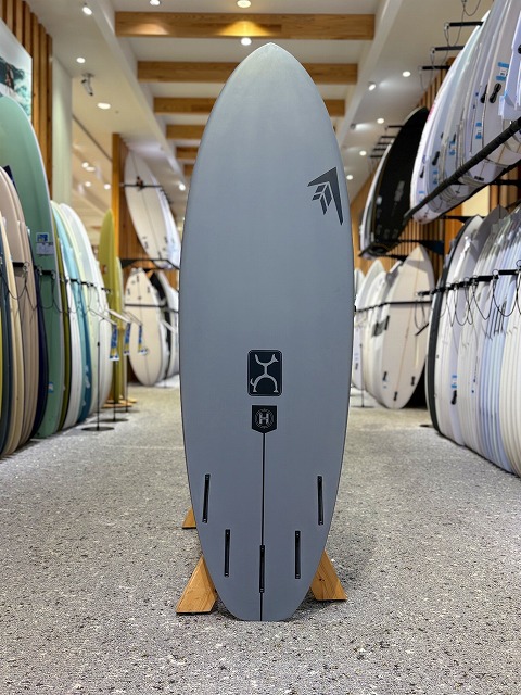 a-image3 - SHOP NEWS | ムラサキスポーツオンラインストア 通販 Gray surfboard displayed upright on a wooden stand in a surf shop showroom, with fin slots and brand logos visible.