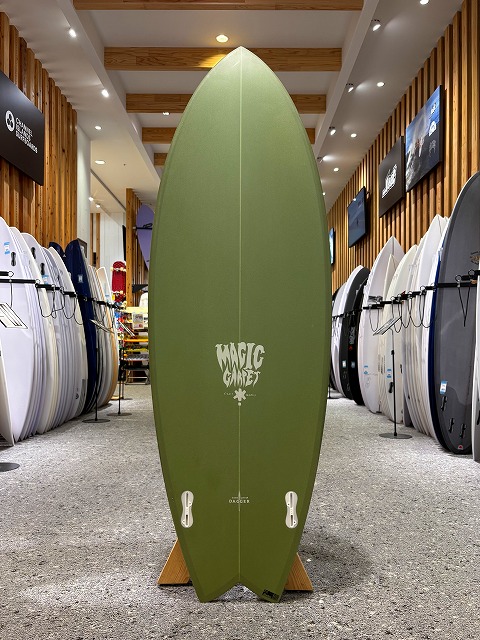 Olive green surfboard standing upright on a display rack in a shop, with rows of surfboards lined up along both walls.
