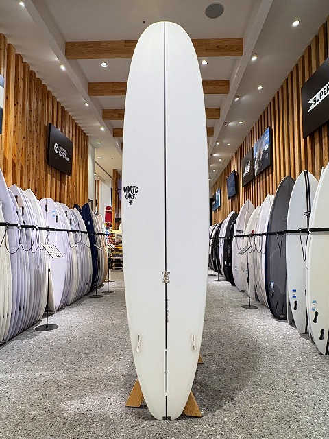 Tall white surfboard standing upright on a wooden stand in a surf shop, surrounded by rows of boards.
