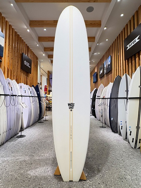 Central white surfboard on display in a shop, indoors with wooden walls and other boards lined up on both sides.